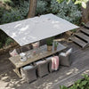 Outdoor patio dining set with a large square white patio umbrella, a wooden dining table, and four woven gray armchairs on a wooden deck surrounded by green plants.