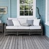 A three-seat outdoor wicker sofa with white and gray cushions, set on a weathered wooden deck in front of a light blue shiplap wall with a white window.