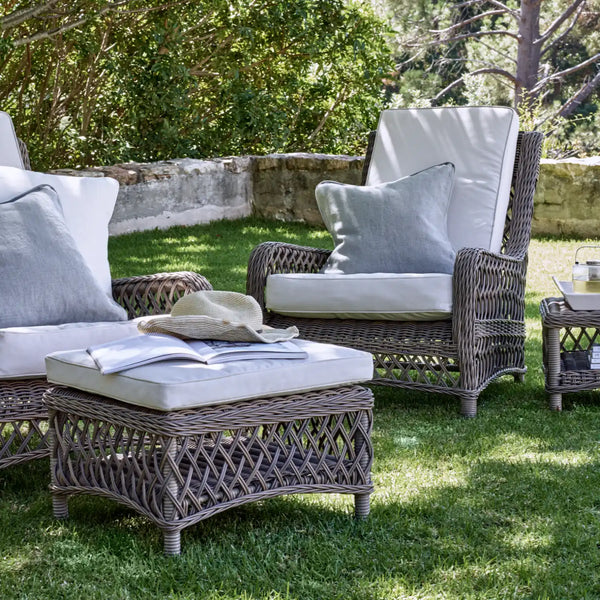 Outdoor patio furniture set on a grassy lawn, featuring a wicker coffee table with a cream cushion, two matching wicker armchairs with light gray cushions, and a small side table in a garden setting.