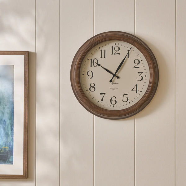 A round wall clock with a dark brown wood-look frame and a cream-colored face, mounted on a beige vertical wood-paneled wall next to a framed picture.