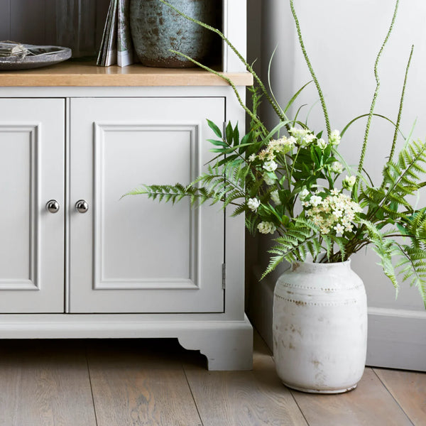 A white cabinet with a light wood top and silver knobs stands against a white wall, with a large white ceramic vase filled with green ferns and small white flowers placed on the floor beside it.