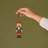 A hand holding a small, colorful, beaded nutcracker ornament against a solid olive green background.