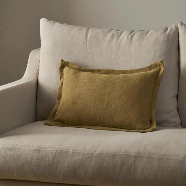 A rectangular saffron yellow linen cushion resting on a light beige upholstered armchair against a neutral gray wall.
