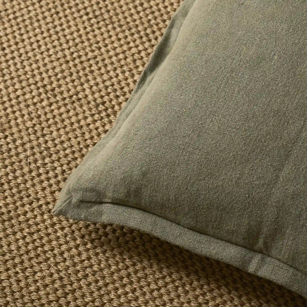 A close-up of a square throw pillow in a muted sage green fabric, resting on a textured beige woven rug.