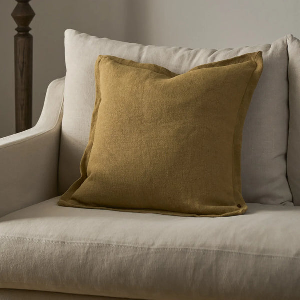 A square mustard yellow linen cushion resting on a light beige upholstered armchair, with a larger off-white pillow behind it.
