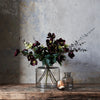 A floral arrangement of dark burgundy hollyhocks, green leaves, and eucalyptus stems in a clear glass jar vase, displayed on a rustic wooden table against a textured gray wall.