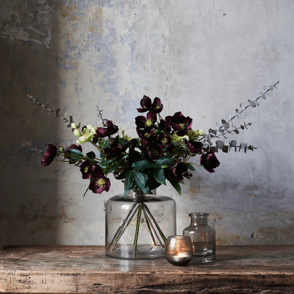A floral arrangement of dark burgundy hollyhocks, green leaves, and eucalyptus stems in a clear glass jar vase, displayed on a rustic wooden table against a textured gray wall.
