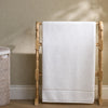 A white bath towel with a folded bottom border draped over a light natural wood towel rack, set against a textured beige wall with soft leaf shadows.