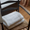 A folded stack of soft white bath towels, placed on the woven seat of a dark wooden chair in a warm indoor setting with wood flooring and curtains in the background.