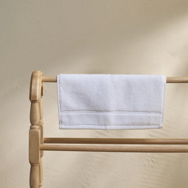 A white cotton hand towel with a stitched border draped over a light natural wood towel rack against a textured beige wall.
