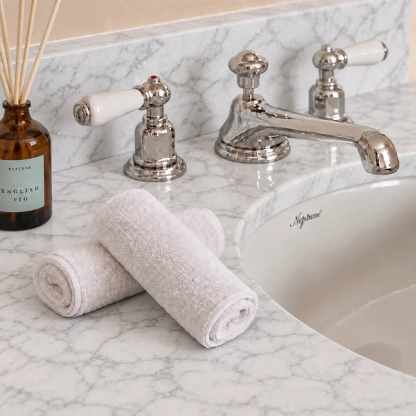 Two rolled white flannels placed on a marble bathroom counter beside a chrome faucet and sink, with a glass reed diffuser visible at left.