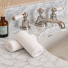 Two rolled white towels on a white marble bathroom counter beside a polished chrome faucet and a small amber reed diffuser bottle.