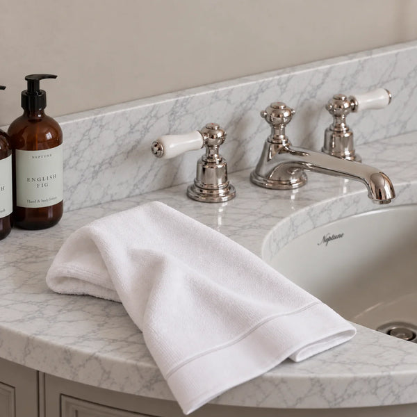 A folded white cotton guest towel with a stitched border rests on a marble bathroom countertop beside two amber pump bottles labeled English Fig and a polished chrome faucet.