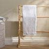 A white cotton hand towel with a folded hem is draped over a light natural wood towel rack with turned spindles, set against a textured beige wall and light wood floor.