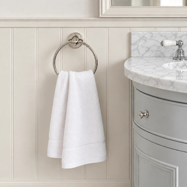 A white hand towel hangs from a round silver towel ring mounted on a cream paneled bathroom wall, with part of a gray vanity and marble countertop visible on the right.