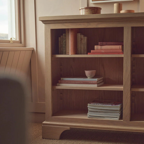 A light oak wooden bookcase with three open shelves, styled with books and a small bowl in a warm, sunlit room.