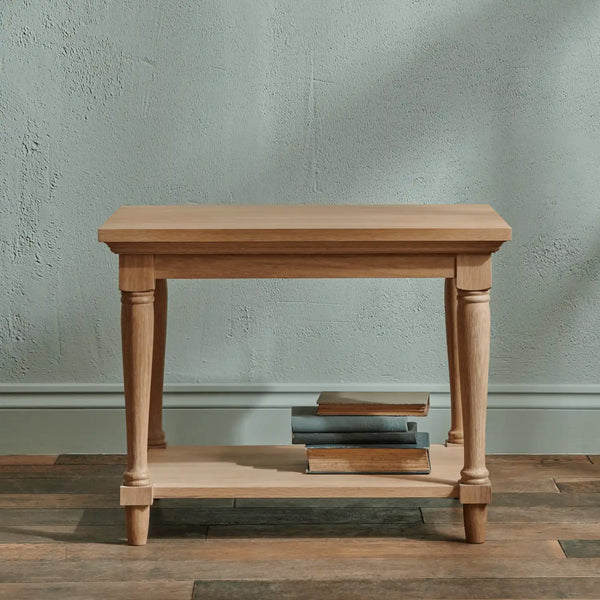 A light natural wood console table with a single shelf, shown on a rustic wood floor against a textured sage green wall. A stack of three books sits on the lower shelf.