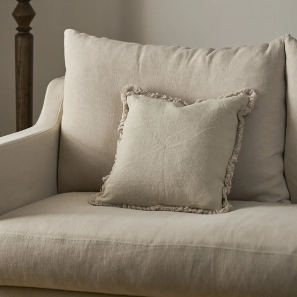 A square beige throw pillow with a fringed edge resting on a matching upholstered armchair in a softly lit interior setting.