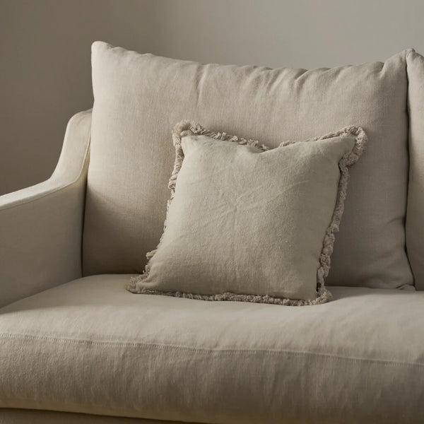 A square beige throw pillow with a fringed edge resting on a matching beige upholstered armchair against a neutral wall.