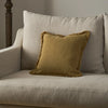 A square yellow throw pillow with a fringed edge resting on a beige upholstered armchair in a softly lit interior setting.