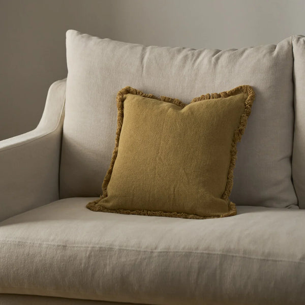 A square yellow throw pillow with a fringed edge resting on a matching beige upholstered armchair against a neutral wall.