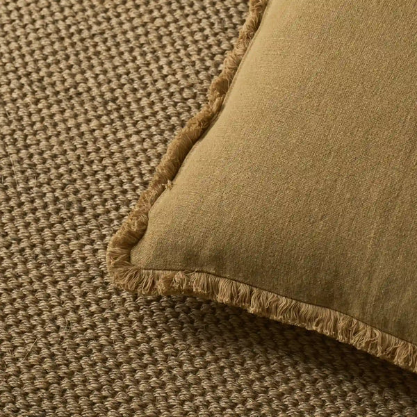 A close-up of a saffron yellow linen throw pillow with fringed edges, resting on a textured brown woven rug.