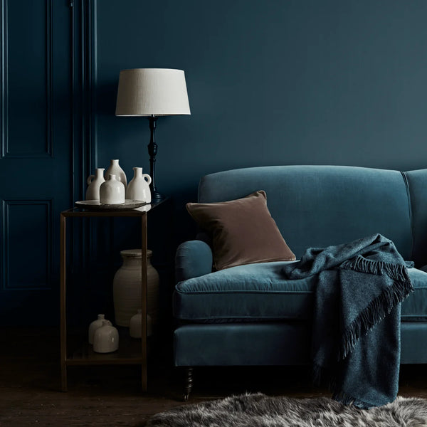 A styled interior scene with a deep teal-blue velvet sofa, a brown throw pillow, a dark blue fringed throw blanket, a black floor lamp with a white drum shade, and a small wooden side table holding white ceramic vases against a dark blue paneled wall.