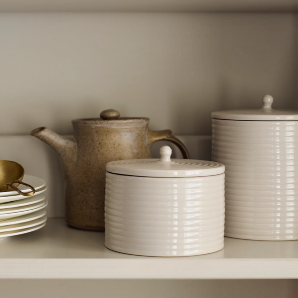 A set of ribbed ceramic canisters in cream and off-white, displayed on a shelf next to a speckled brown teapot and stacked white plates.