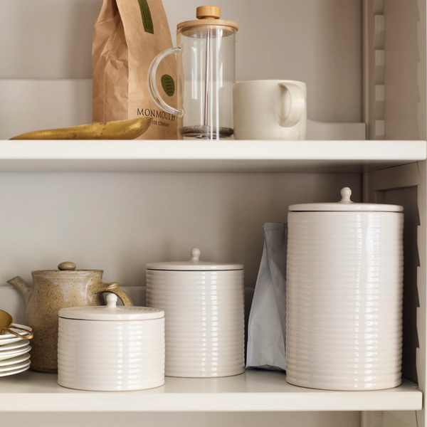 A set of ribbed ceramic canisters in warm off-white, styled on white kitchen shelves alongside other home goods including a glass carafe, a brown paper bag, and a ceramic mug.