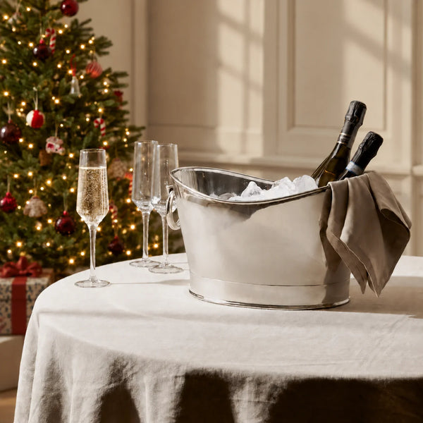 A silver metal ice bucket filled with ice and two wine bottles, set on a white tablecloth next to two champagne flutes. A decorated Christmas tree with warm lights is blurred in the background.