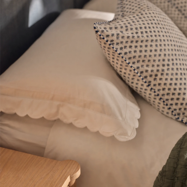 Close-up of a bed with cream-colored bedding and two pillows, including one with a small dark geometric pattern and scalloped edge.
