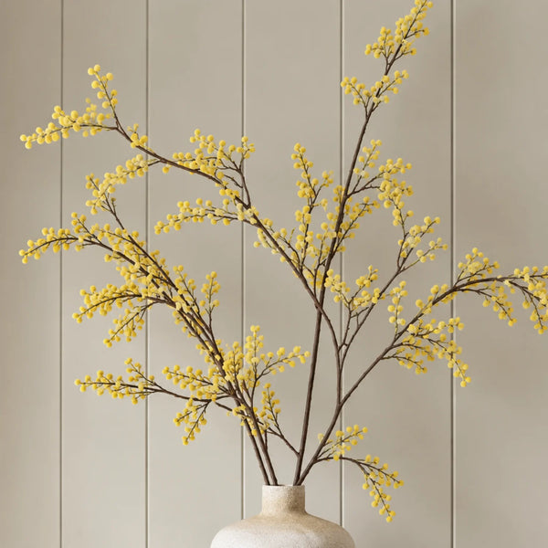 Yellow flowering branches arranged in a small cream-colored ceramic vase against a beige paneled wall background.