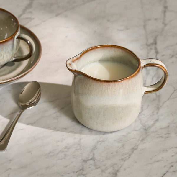Cream ceramic milk jug with brown glazed rim on a marble tabletop beside a spoon and cup.