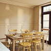 A dining room scene with a light wood rectangular table surrounded by six yellow spindle-back chairs, set with plates and glasses near a large dark-framed window.