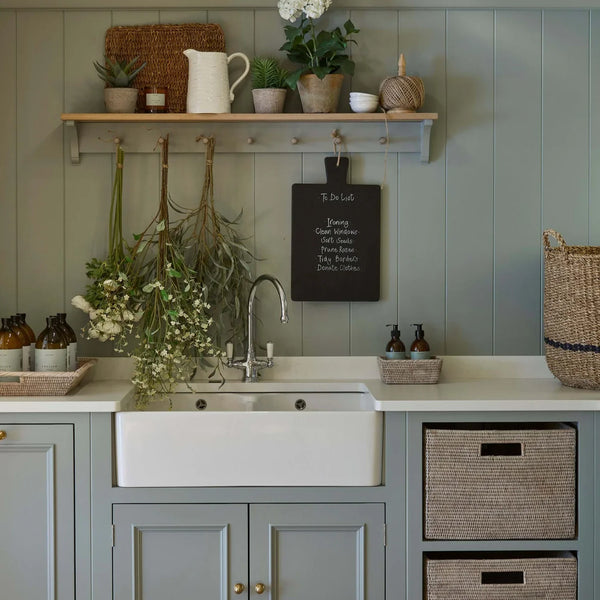 Utility room with sage green panelled walls, farmhouse sink, open shelf and woven storage baskets.