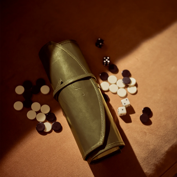 A rolled olive green leather game bag lies on a warm brown surface, surrounded by scattered black and white game pieces and dice under dramatic directional lighting.