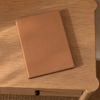 A tan leather notebook with stitched edges and a small embossed monogram, resting on a light wood table.