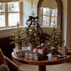 Four small artificial Christmas trees in woven planters arranged on a round wooden table, with wrapped gift boxes and candles in a warm, decorated indoor room.