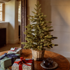 A small artificial Christmas tree in a woven basket pot, lit with warm white string lights, set on a wooden table next to wrapped gift boxes in a cozy indoor room.