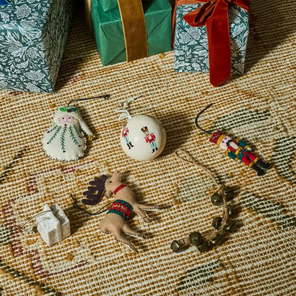 A styled flat-lay of several handmade Christmas ornaments on a woven beige rug, with wrapped gift boxes in the background. The ornaments include a white angel with green details, a white nutcracker ball, a colorful wooden nutcracker, a small plush reindeer, and a rustic jingle bell garland.