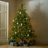 A decorated artificial Christmas tree with warm white lights stands in a corner of a room, with wrapped gift boxes at its base. The tree is adorned with white ornaments, small hanging figures, and rustic wooden accents.
