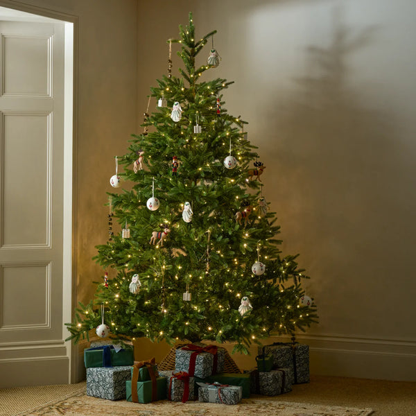 A decorated artificial Christmas tree with warm white lights stands in a corner of a room, with wrapped gift boxes at its base. The tree is adorned with white ornaments, small hanging figures, and rustic wooden accents.