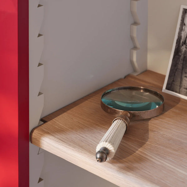 A vintage-style magnifying glass with a ribbed cream handle and a bronze metal rim rests on a light wood shelf next to a white slatted wall panel.