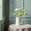 White ranunculus faux flower bundle shown in a white stoneware jug on a beige linen cloth covered table against a blue wall