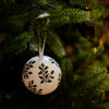 A white spherical Christmas ornament decorated with green holly leaves and small red berries, hanging from a green Christmas tree branch.