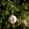 A white Christmas ornament shaped like a nutcracker soldier hanging from a green Christmas tree with warm string lights.