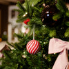 A close-up of a decorated Christmas tree with green pine branches, a red and white striped hanging ornament, a glossy black ornament, warm string lights, and a pink striped bow.