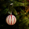 A round Christmas ornament with red and white vertical stripes hanging from a green Christmas tree branch.