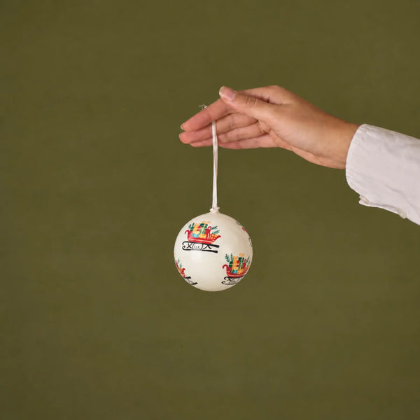 A hand holding a small white Christmas ornament with colorful sleigh illustrations against a solid olive green background.