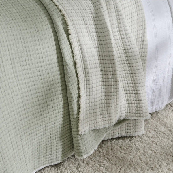 A close-up of a pale sage green waffle-weave throw blanket draped over a bed with white bedding and a beige carpet visible underneath.